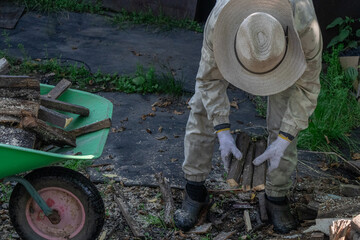 A beekeeper in protective gear loads firewood into a wheelbarrow in a rural setting.
