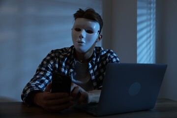 Anonymity. Masked man with smartphone and laptop at table in dark