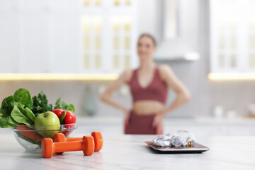 Slim woman in sportswear posing indoors, focus on kitchen scale, healthy food, dumbbells and...