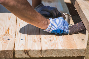 A carpenter wearing cloth gloves manually polishes wooden boards with sandpaper for grinding. The...