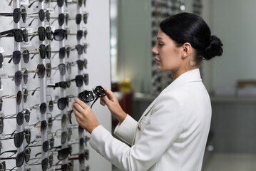 Attractive Caucasian young brunette woman in white suit choosing sunglasses on showcase for purchase at an optical store. Lifestyle, optometry, eyesight, assortment of glasses