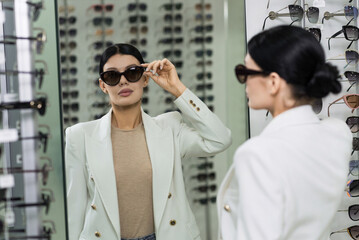 Elegant Caucasian young brunette woman in white suit trying on sunglasses looking in mirror for purchase at optical store. Lifestyle, optometry, eyesight, eye protection