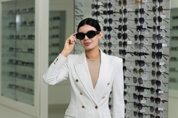 Attractive Caucasian young brunette woman in white suit trying on and choosing sunglasses to buy at an optical store. Lifestyle, optometry, eyesight, assortment of glasses