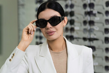 Attractive Caucasian young brunette woman in white suit trying on and choosing sunglasses to buy at an optical store. Lifestyle, optometry, eyesight, assortment of glasses.
