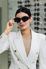 Attractive Caucasian young brunette woman in white suit trying on and choosing sunglasses to buy at an optical store. Lifestyle, optometry, eyesight, assortment of glasses.