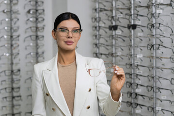 Attractive Caucasian young brunette woman in white suit trying on and choosing eyeglasses for buying at an optical store. Lifestyle, optometry, eyesight, assortment of glasses