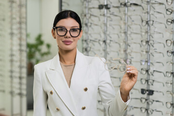 Attractive Caucasian young brunette woman in white suit trying on and choosing eyeglasses for buying at an optical store. Lifestyle, optometry, eyesight, assortment of glasses