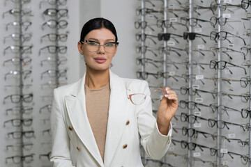 Attractive Caucasian young brunette woman in white suit trying on and choosing eyeglasses for buying at an optical store. Lifestyle, optometry, eyesight, assortment of glasses