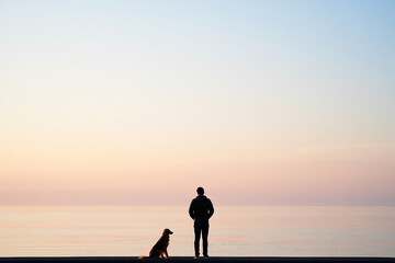 Fototapeta premium Silhouetted figures of a man and his dog watch a serene sunrise over the ocean