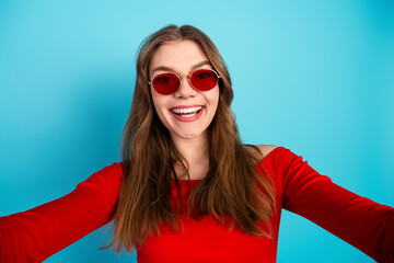 Stylish young woman in red top, trendy sunglasses, smiling with confidence against vibrant blue...