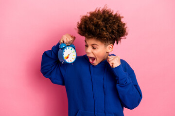 Excited boy holding a clock and wearing a bright blue hoodie against a vibrant pink background,...