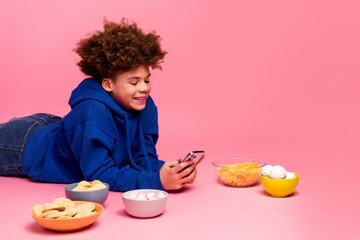 Young boy in blue hoodie enjoying snacks and using smartphone on pink background, representing...