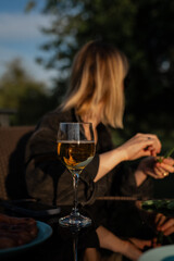 Glass of white wine on reflective table with woman sitting outdoors in warm evening light enjoying relaxing moment in garden with nature background and casual atmosphere