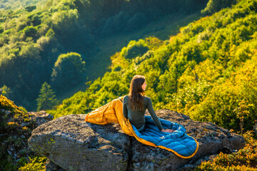 Woman sitting on a cliff in a sleeping bag, watching sunrise over forested mountains