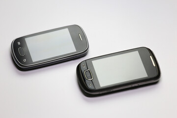 A close-up perspective of two vintage mobile phones, featuring their front faces with grey screens,...