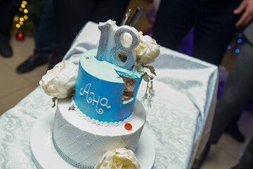 A two-tiered blue and white birthday cake decorated with a pearl ribbon and white and yellow flowers with the female name Ana written in Cyrillic script