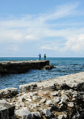 Two men fishing from a rocky pier stretching into the sea under a blue sky with scattered clouds. Peaceful coastal scene with natural textures and relaxing atmosphere. Outdoor leisure concept.