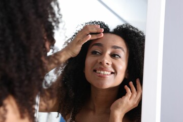 Beautiful Brazilian woman with healthy skin looking at mirror indoors
