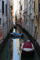  Boats with colorful covers are moored in a quiet, narrow canal, with their reflections perfectly mirrored in the dark, still water.
