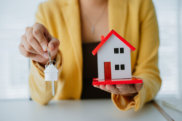 Close-up of hand presenting house-shaped keychain over mortgage papers and model house, symbolizing...