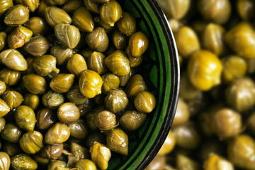 Capers in a round bowl. Pickled buds with greenish color and wrinkled texture. Salty flavor used in...