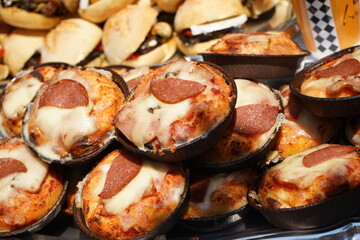 Mini pizzas are displayed outdoors in black baking trays, topped with melted cheese and pepperoni slices. The crust is golden brown, and the cheese is slightly browned as they are freshly baked