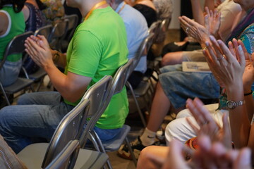 A diverse audience seated on folding chairs claps in applause. Casual attire like t-shirts and jeans is visible. Focus on clapping hands, with a notebook in view, in a modern conference setting.