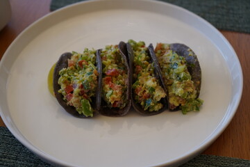 Four mini tacos with an avocado-based filling rest on a white plate. Made with blue corn tortillas,...