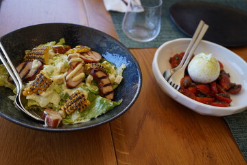 Fresh Caesar salad with grilled chicken, charred corn, romaine lettuce, croutons, and creamy dressing, alongside Mozzarella cheese with cherry tomatoes and olives. Elegant dining in a Greek hotel.