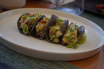 Four mini tacos with an avocado-based filling rest on a white plate. Made with blue corn tortillas, they feature diced tomatoes and herbs, offering a fresh, colorful touch on a wooden table.