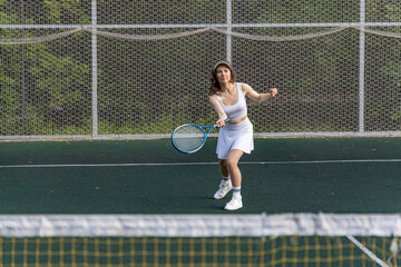 Woman in white tennis outfit holding a blue racket, standing on a green clay court, ready to serve...