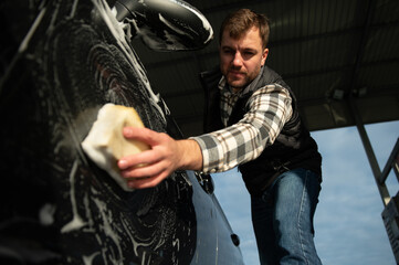 Man worker with a sponge washes a car with foam. Car wash and car care
