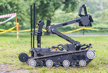 A multi-wheeled, black and silver robotic device is depicted on a grassy area with trees in the background. The robot, resembling heavy machinery, has sensors or cameras