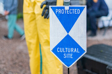 Two individuals in bright yellow rain gear stand near a blue signpost with CULTURAL SITE written on it, possibly under construction or maintenance, in an outdoor setting under an overcast sky