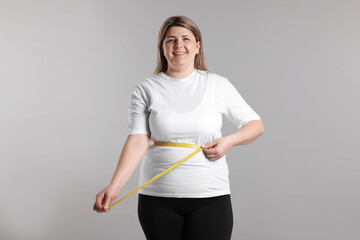 Weight loss. Plus size woman measuring waist with tape on gray background