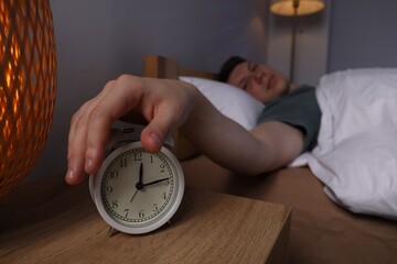 Frustrated man suffering from insomnia and turning off alarm clock in bed indoors, selective focus