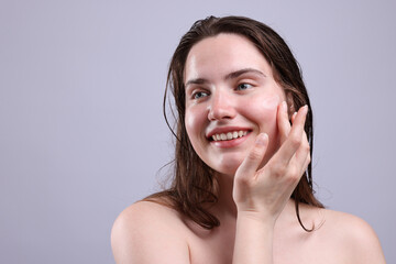 Smiling woman applying skincare product onto her face on grey background, closeup. Space for text