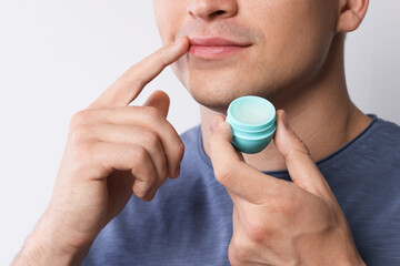 Young man applying lip balm onto his lips on light grey background, closeup