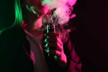 Woman using electronic smoking device in neon lights on black background, closeup. Space for text