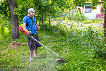 Senior man trimming overgrown backyard with strimmer. Older man wearing safety goggles uses a red...