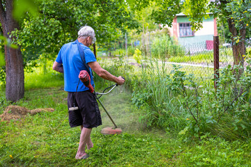 Senior man cutting tall grass with string trimmer. Older man in blue shirt uses a red string trimmer to cut overgrown grass along a fence in a leafy backyard garden.
