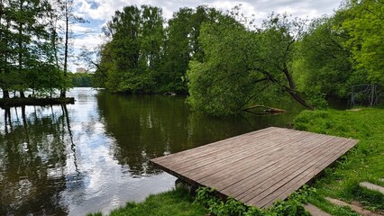 A wooden pier and a leaning willow tree stand on the shore of the lake. Nearby is a metal fence. The sky and trees are reflected in the water. On the far shore is a park with trees and buildings