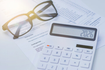 White calculator displaying the number 2026, placed on financial documents with black eyeglasses nearby. Concept business planning and tax calculation.