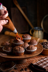 Chocolate muffins with liquid chocolate and dulce de leche with cocoa powder, a chocolate bar, and coffee beans on a wooden table with kitchen utensils.