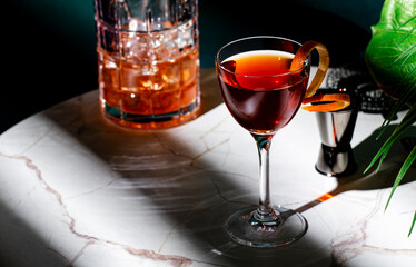 beautiful crafted cocktail in glass garnished with orange peel on marble table beside an ice-filled glass and bar tools.