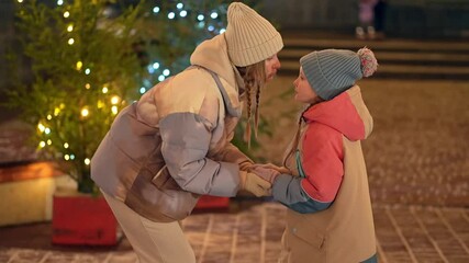 Caucasian mother is kissing her little daughter standing holding hands on the street near decorated trees. Slowmo of woman and child in hats and jackets spending winter evening in Christmas fairground - Powered by Adobe