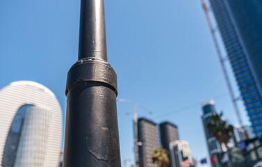 A minimalist city scene captured with foreground focus on a black metal pole against blurred modern skyscrapers and a clear blue sky. Perfect for design projects exploring urban life. Selective focus.