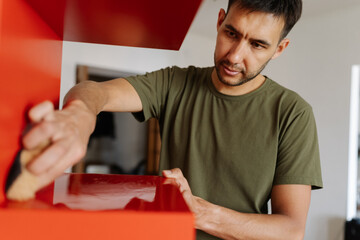 Portrait of young focused man cleaning wiping red shelf surface with sponge and detergent ensuring...