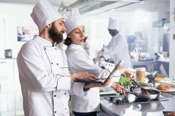 Team of experts with hats researching new meal recipes on a laptop before preparing ingredients,...