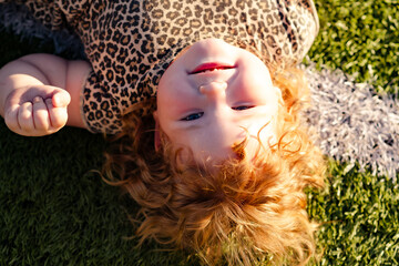 A young child with curly red hair is lying on green grass smiling and playing happily under the...